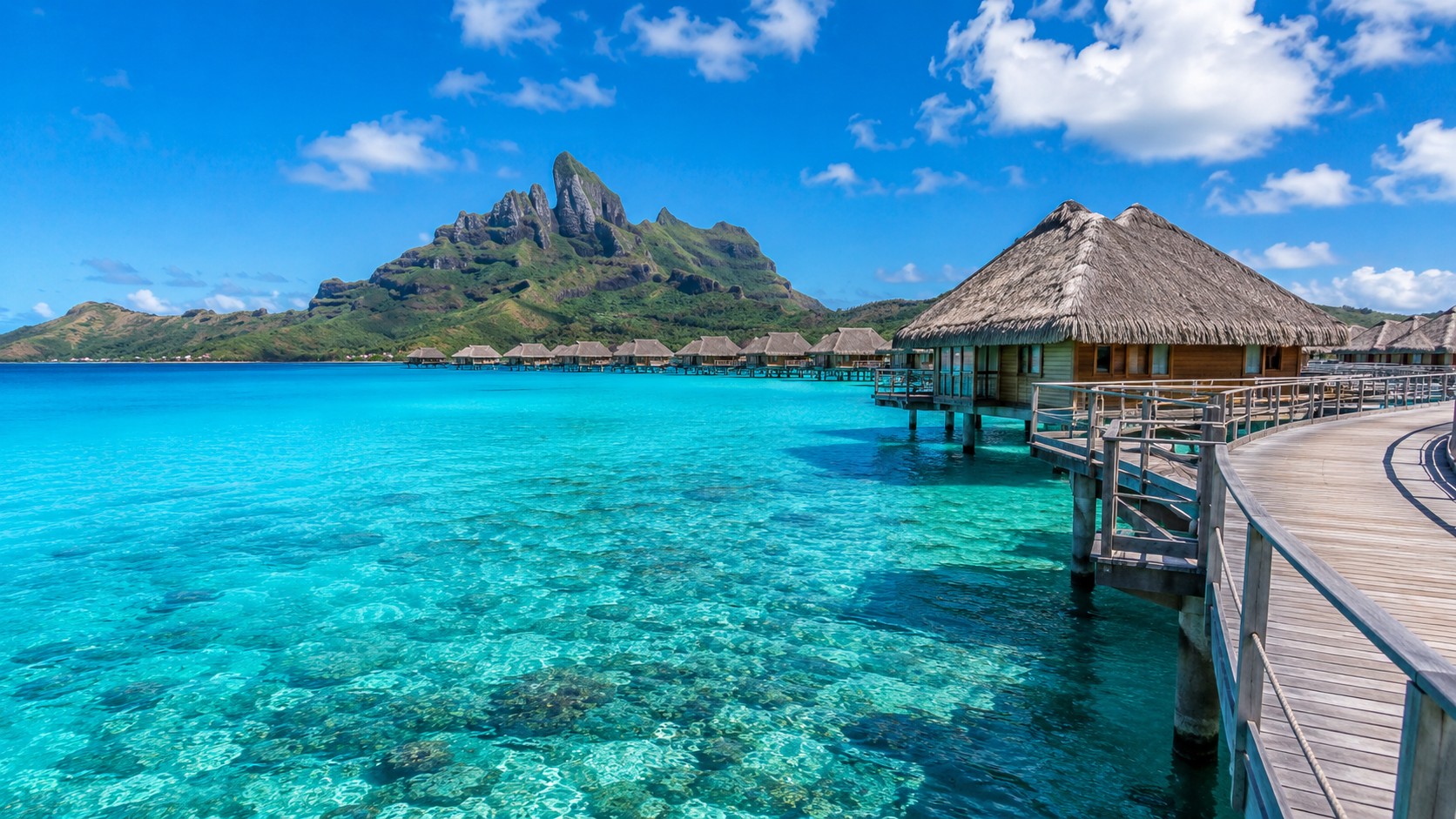 Photo professionnelle ultra réaliste format 16/9 d’un paysage tropical dans l’océan Pacifique avec bungalows sur l’eau, lagon turquoise, ponton en bois, montagnes verdoyantes et ciel ensoleillé.