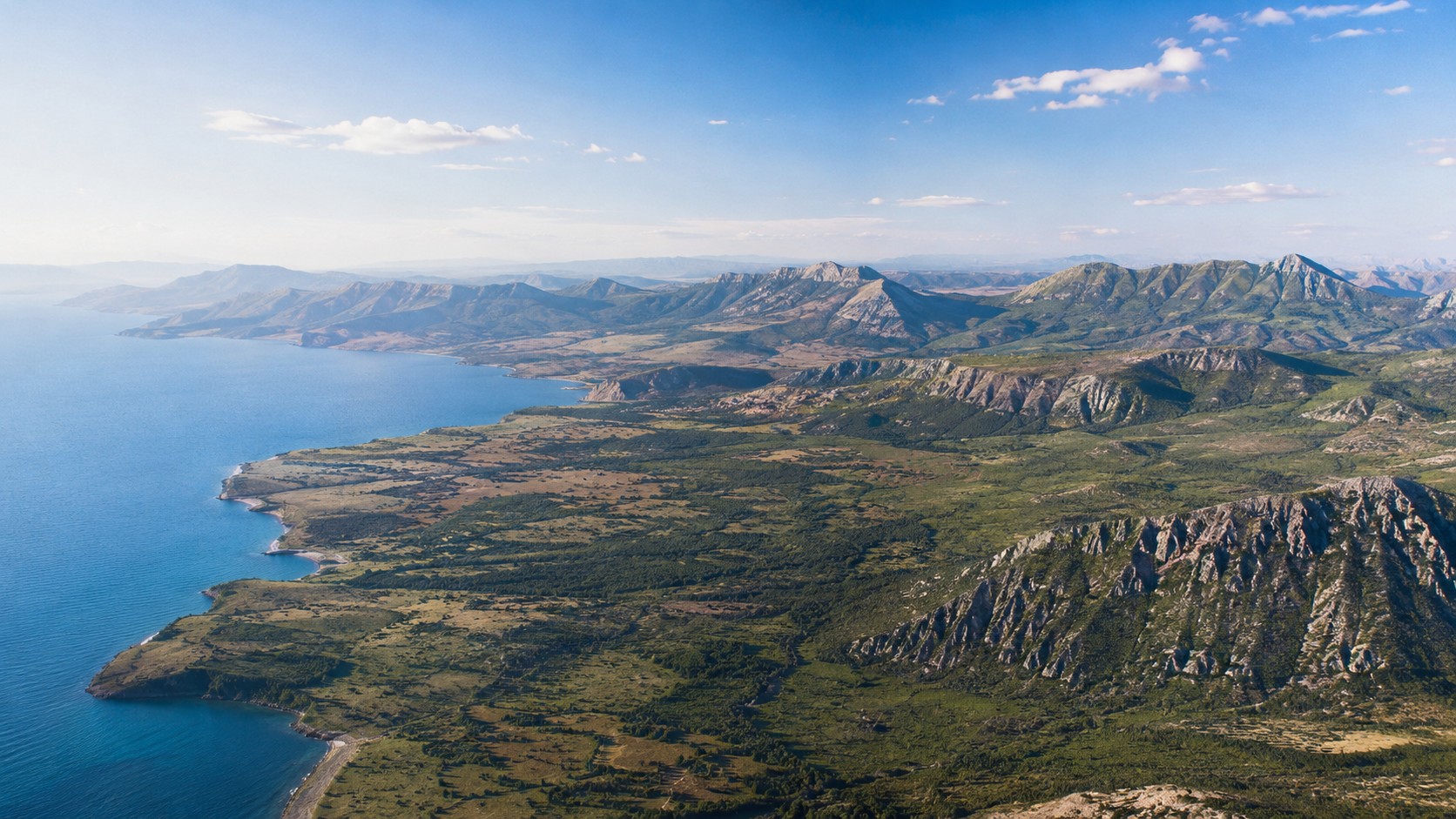 Photo professionnelle ultra réaliste en format 16/9 d’un vaste continent vu en vue aérienne, avec littoral bleu, plaines verdoyantes, reliefs rocheux, montagnes lointaines et ciel ensoleillé, style banque d’images premium naturel et éditorial