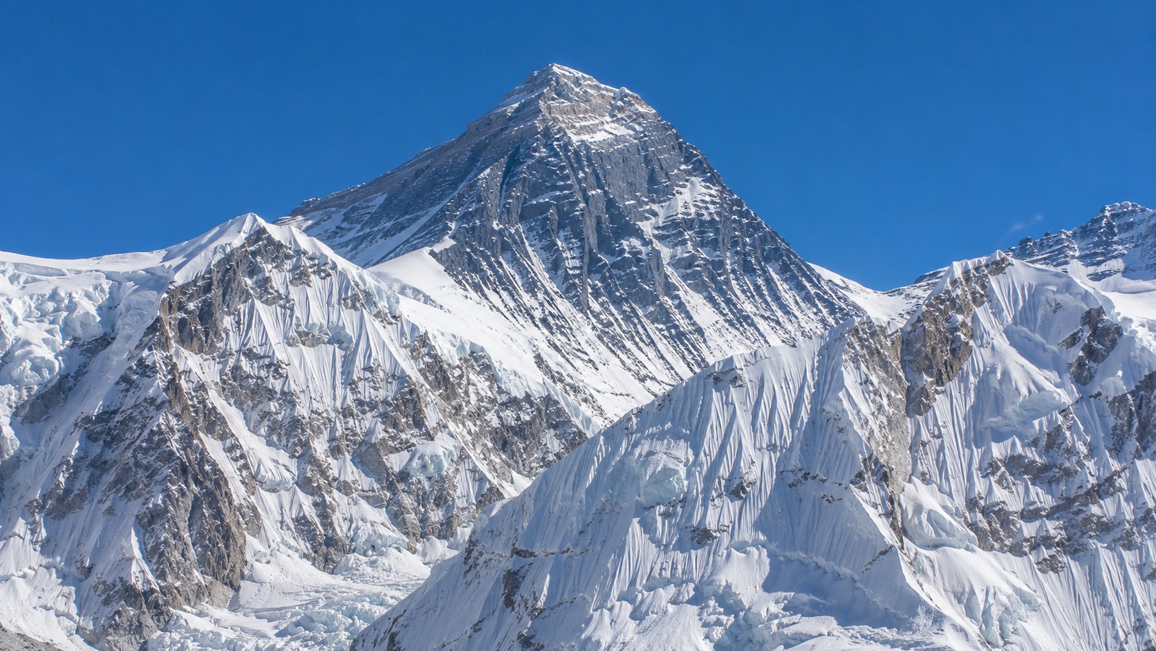 Photo professionnelle ultra réaliste de l’Everest enneigé dans l’Himalaya, avec sommet majestueux, crêtes glacées, textures de neige et de roche détaillées, ciel bleu clair et météo ensoleillée, format 16/9.