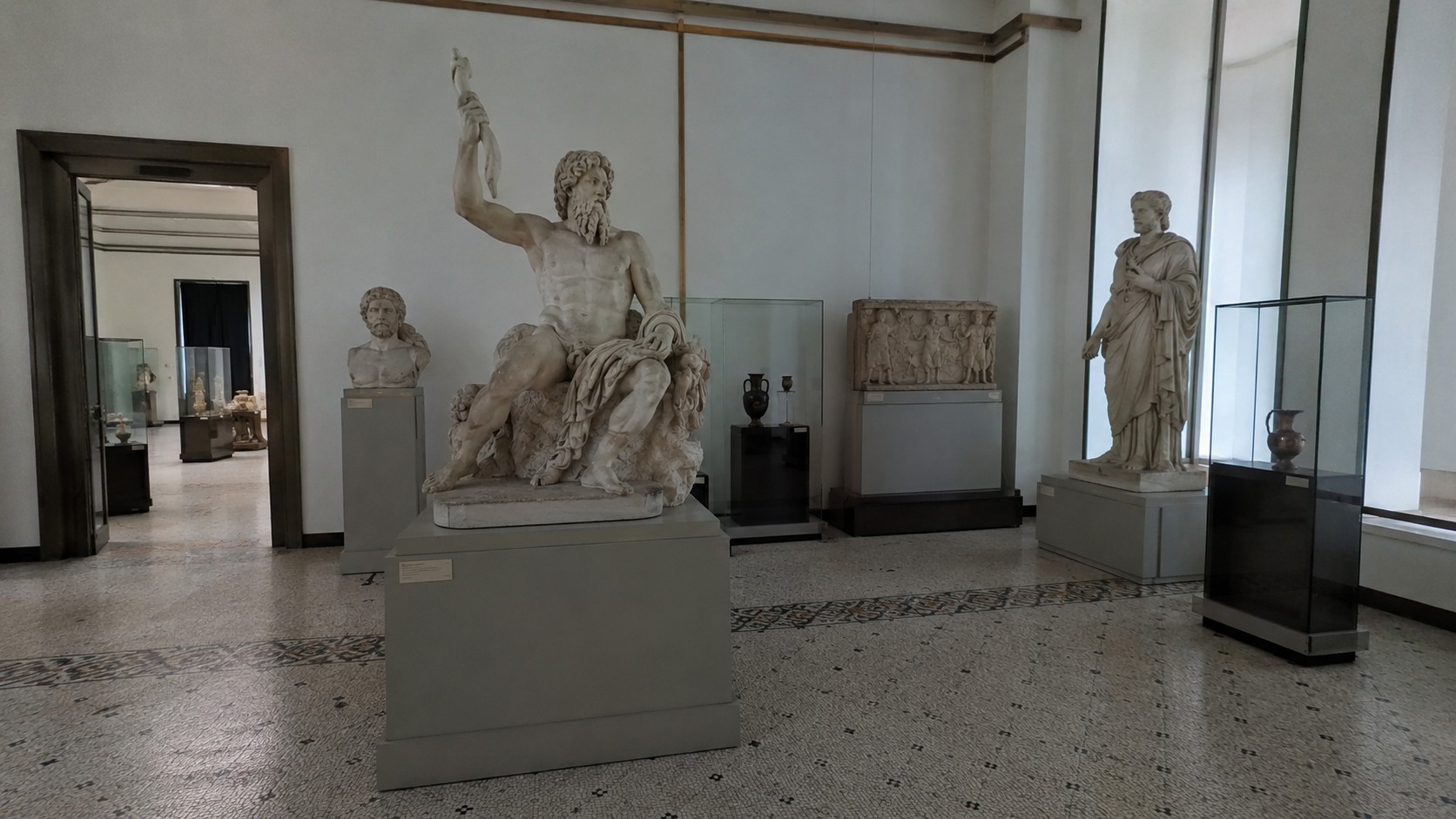 Cette photographie de style documentaire, réalisée comme avec un Panasonic Lumix GH4, présente une collection de statues antiques inspirées de la mythologie grecque. On y distingue des représentations des grands dieux olympiens tels que Zeus, Athéna, Poséidon ou Apollon, ainsi que plusieurs héros légendaires comme Héraclès ou Persée. La lumière naturelle met en valeur les détails du marbre, les textures vieillies des sculptures et l'architecture classique du lieu. L'image évoque les récits fondateurs de la Grèce antique, les mythes, les légendes et l'héritage culturel grec dans un rendu réaliste et authentique.