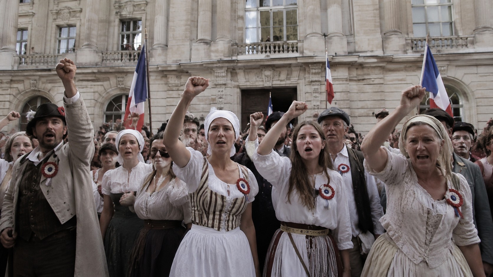 Foule de citoyens et citoyennes en tenue d’époque de la Révolution française de 1789, brandissant le poing et des drapeaux tricolores devant un bâtiment historique en pierre à Paris. Scène de reconstitution historique montrant l’engagement populaire, les cocardes révolutionnaires et l’atmosphère des événements ayant marqué la chute de l’Ancien Régime en France.